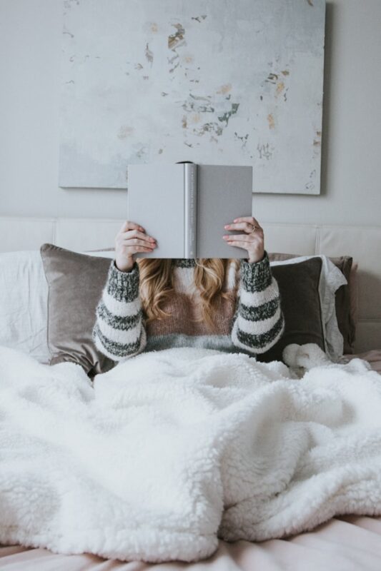 girl in striped sweater reading a gray book on bed covered by white cozy blanket